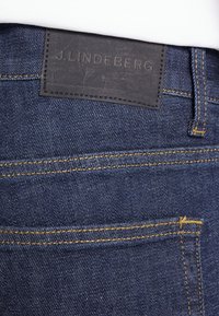 Mörkblå jeans i denim med en läderlapp märkt "J.Lindeberg" på bakre midjan, med gul söm längs sömmarna.