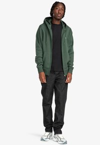 Sudadera verde con capucha y bolsillos frontales, combinada con una camiseta negra, jeans oscuros y zapatillas deportivas blancas/grises.