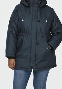 Marinblå vadderad jacka med huva, tryckknapplikationer och knappstängning, elastisk midja och sidofickor. Slät textur med en strukturerad passform.