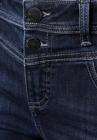 Jeans en denim bleu foncé avec des coutures argentées et une texture lisse. Comprend deux boutons noirs avec un logo et une petite poche avant.