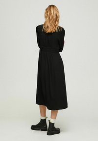 Vestito nero con vita stretta, lunghezza al ginocchio, maniche lunghe e una texture liscia. Abbinato a stivali neri alla caviglia e calzini bianchi.