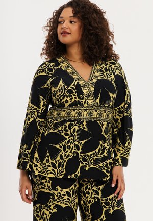 Frau trägt ein gelb-schwarzes Oberteil mit V-Ausschnitt, bedruckt mit Blumen und Vögeln, und passende weite Hosen, steht vor einem weißen Hintergrund.