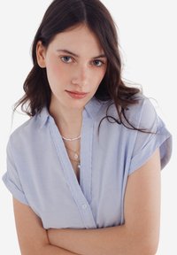 Camicia a righe azzurre con maniche corte, texture morbida, colletto, chiusa con bottoni, indossata con collane a più strati.