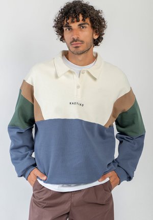 Hombre con pelo rizado que lleva un jersey con bloques de color en crema, marrón, verde y azul, de pie con las manos en los bolsillos.