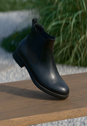 Bottine en cuir noir avec des panneaux élastiques sur les côtés, bout rond et talon bas, exposée sur une surface en bois contre un décor verdoyant.