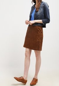 Gonna di camoscio marrone abbinata a una maglietta blu e a un giubbotto di jeans scuro. Scarpe in pelle marrone completano l'outfit, mostrando un look casual.