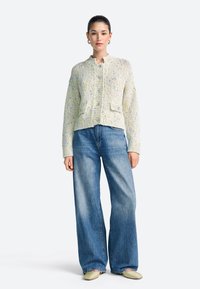 Teksturiran svetlo rumen kardigan z okroglim izrezom in dvema sprednjima žepoma, kombiniran z modrimi kavbojkami širokih hlačnic in svetlimi čevlji.