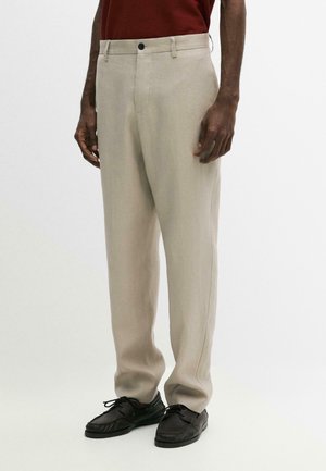 Mann trägt beige gerade geschnittene Hose, dunkelrotes Oberteil und schwarze Leder-Schnürschuhe, steht vor einem einfarbig weißen Hintergrund.