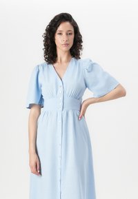 Rochie cămașă de culoare albastru deschis, cu mâneci scurte tip puff. Are o talie ajustată și o fustă fluidă, confecționată dintr-un material texturat.
