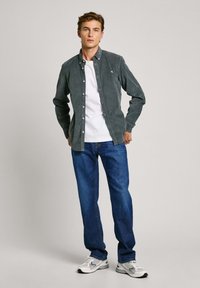 Grijze corduroy knoopshirt over een witte T-shirt, gecombineerd met blauwe jeans en grijze sneakers. Staande model tegen een lichtgrijze achtergrond.