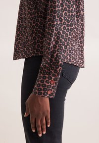 Maglietta tampon in tessuto rosso scuro con motivo floreale nero. Maniche a cuffia con due bottoni rotondi, abbinata a jeans scuri.