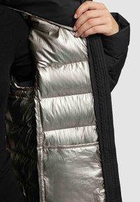 Zwarte pufferjack met een glanzende metallic zilveren voering, voorzien van de gestikte secties en een verborgen zak aan de binnenkant van de linkerzijde.