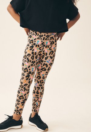 Kind trägt bunte Leggings mit Leopardenmuster und schwarze Sneaker, steht mit den Händen in der Hüfte vor einem schlichten Hintergrund.