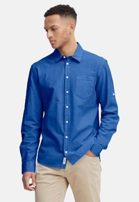 Langärmliges Button-up-Hemd in leuchtendem Blau, aus einem strukturierten Stoff gefertigt. Verfügt über eine Brusttasche und weiße Knöpfe. Kombiniert mit beigen Hosen.