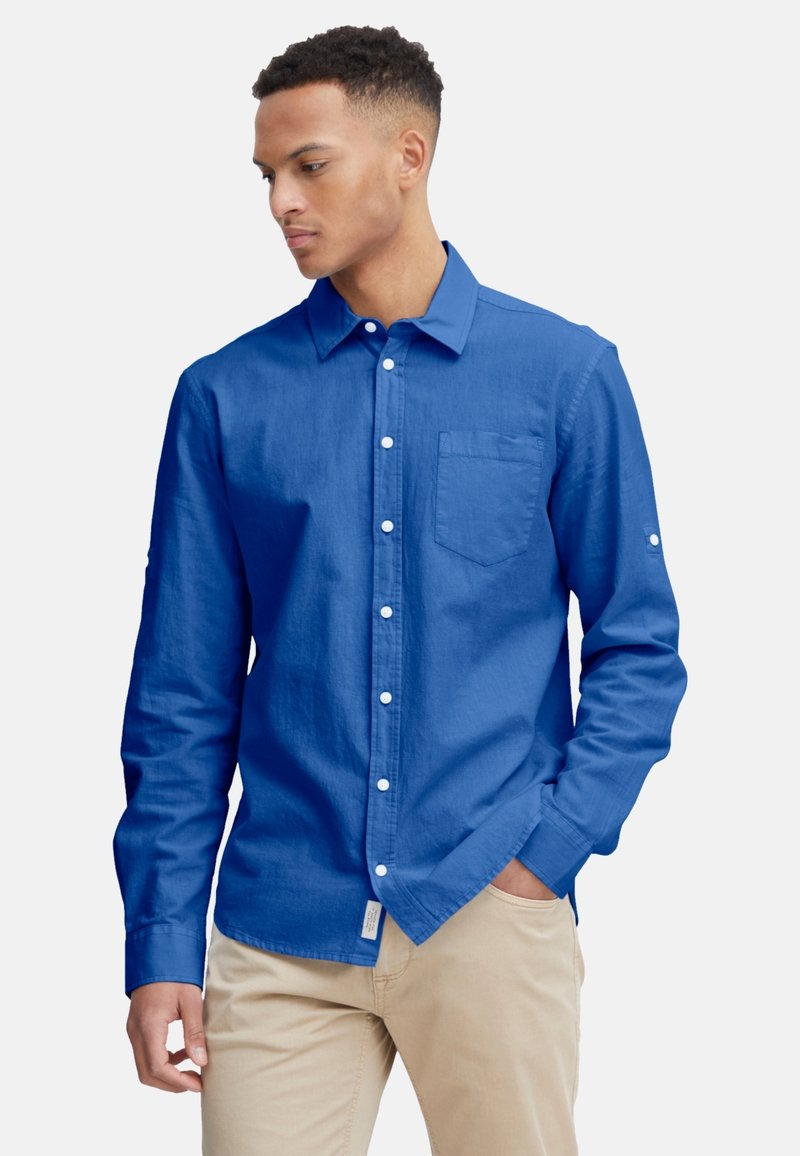 Langärmliges Button-up-Hemd in leuchtendem Blau, aus einem strukturierten Stoff gefertigt. Verfügt über eine Brusttasche und weiße Knöpfe. Kombiniert mit beigen Hosen.