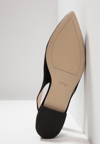 Zapato de tacón alto de ante negro con punta afilada, laterales con cortes, y una suela beige lisa, que presenta el nombre de la marca grabado en la plantilla interior.