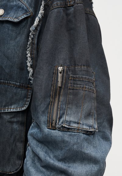 Μανίκι σκουρόχρωμου ξεβαμμένου τζιν μπουφάν με τσεπάκι με φερμουάρ και διπλανή ραμμένη τσέπη σε απλό ανοιχτό φόντο.