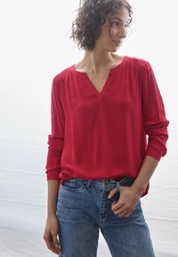 Blusa rossa a scollo a V con spalle arricciate, maniche lunghe e tessuto morbido, abbinata a jeans in denim azzurro chiaro. Sfondo semplice.