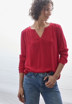 Blouse rouge à col en V avec des épaules froncées, manches longues et tissu doux, associée à un jean en denim bleu clair. Fond uni.