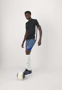 T-shirt de sport noir Nike avec des accents blancs, shorts gris avec des détails bleus, chaussures noires et chaussettes blanches, debout sur un ballon de soccer.