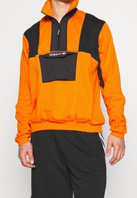 Felpa con cerniera arancione e nera con collo alto, dotata di una tasca frontale e del logo Adidas sul petto. Tessuto morbido al tatto.