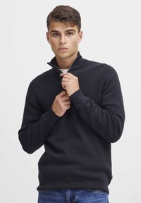 Schwarzer Pullover mit Reißverschluss im kragenlosen Design aus strukturiertem Strickmaterial, figurnah geschnitten, mit gerippter Kragen- und Bündchenstruktur, getragen über blauen Jeans.