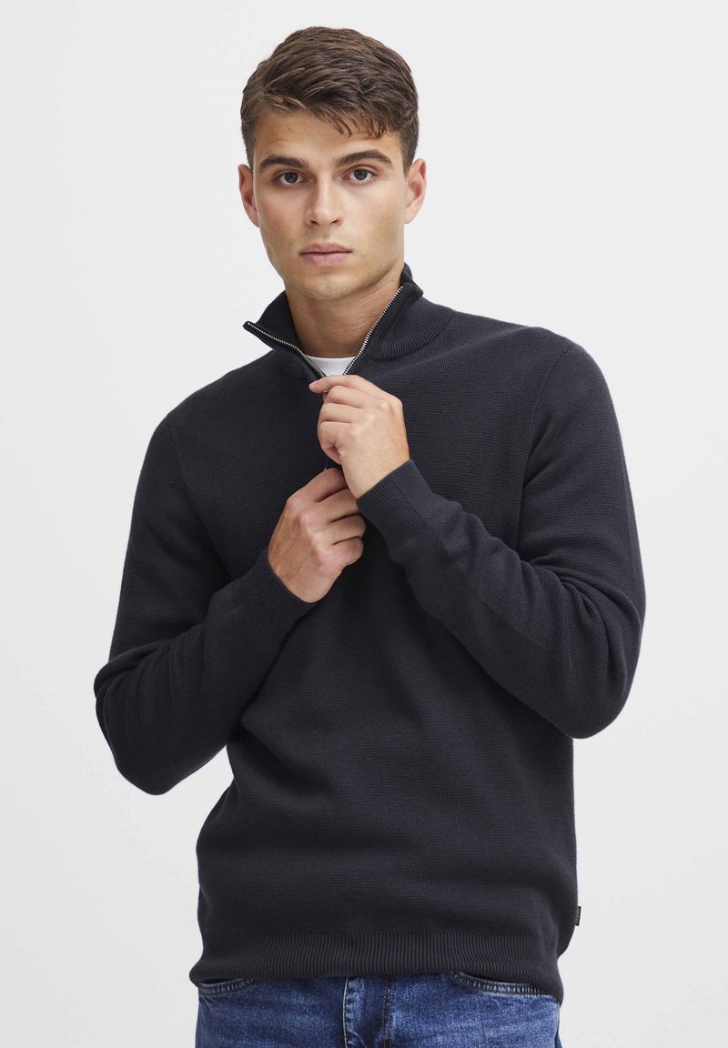 Schwarzer Pullover mit Reißverschluss im kragenlosen Design aus strukturiertem Strickmaterial, figurnah geschnitten, mit gerippter Kragen- und Bündchenstruktur, getragen über blauen Jeans.