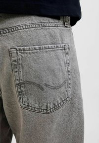 Des jeans en denim gris présentent une poche arrière avec un design de couture contrastant. Le tissu semble texturé, avec un léger dégradé de couleur.
