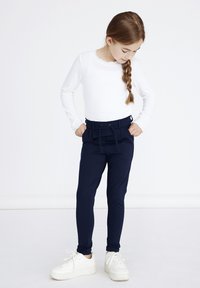 Witte longsleeve top met een geschulpte halslijn, gecombineerd met marineblauwe slim-fit broeken met een elastische taille en zijzakken. Witte sneakers.