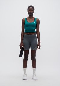 Γυναίκα που στέκεται φορόντας τοπ σε τιρκουάζ χρώμα, γκρι αθλητικά σορτς, λευκά αθλητικά παπούτσια, κρατώντας μαύρο παγούρι νερού μπροστά σε λευκό φόντο.