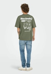 Olijfgroen katoenen t-shirt met een grote witte afbeelding van een vintage scooter en de tekst "Slow Sunday" op de achterkant. Relaxte pasvorm, korte mouwen.