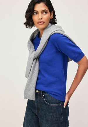 Frau trägt einen königsblauen Kurzarm-Pullover mit einem grauen Pullover über den Schultern und dunkelblaue Jeans, schaut zur Seite.