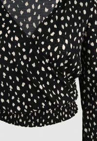 Black fabric top with white irregular polka dots, featuring a wrap design and elastic gathered hem. Textured, lightweight material, long sleeves.