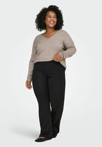 Femme souriante aux cheveux bouclés portant un pull beige à col en V, un pantalon noir et des ballerines noires, se tenant devant un fond blanc.