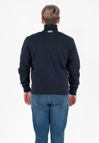 Marineblauwe sweatshirt met hoge kraag, geribbelde onderrand en manchetten, voorzien van een subtiel logo op de achterkant, gecombineerd met blauwe spijkerbroek.