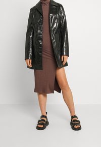 Cappotto in pelle nera lucida con colletto a punte e due tasche frontali, indossato sopra un vestito marrone a coste, abbinato a sandali neri.