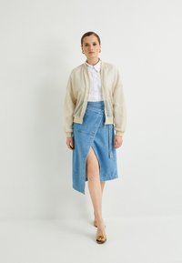 Chaqueta bomber de malla color beige claro sobre una camiseta blanca, combinada con una falda de mezclilla azul con un corte y sandalias de tacón amarillas.