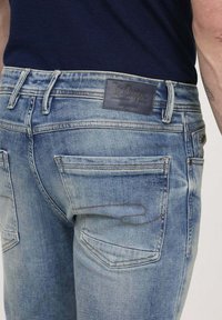 Un jean en denim bleu clair présente une étiquette en cuir marron foncé au niveau de la taille, deux poches arrière avec des coutures contrastées, et un design ajusté.