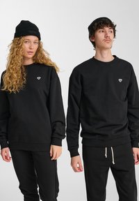 Due persone che indossano berretti di maglia nera, felpe nere con piccoli loghi sul petto e pantaloni neri, in piedi contro uno sfondo bianco semplice.