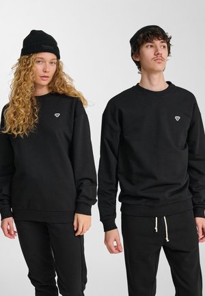 To personer iført sorte strikkede beanies, sorte sweatshirts med små brystlogoer og sorte bukser, står mod en ensfarvet hvid baggrund.