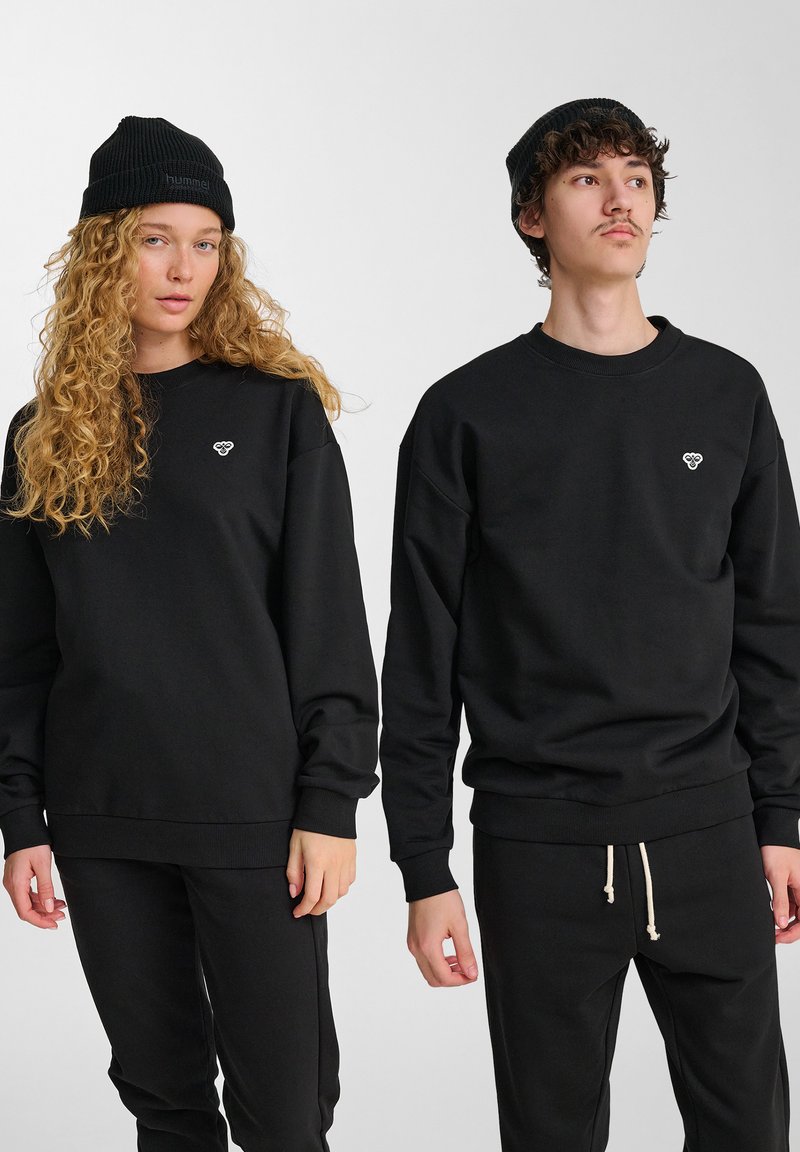 Due persone che indossano berretti di maglia nera, felpe nere con piccoli loghi sul petto e pantaloni neri, in piedi contro uno sfondo bianco semplice.