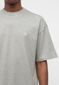 Άτομο που φορά ανοιχτογκρι T-shirt Adidas με μικρό λευκό λογότυπο τριφύλλι στο αριστερό στήθος, κοντά μανίκια και στρογγυλή λαιμόκοψη.