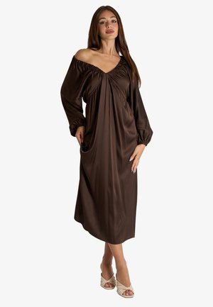Donna che indossa un lungo vestito marrone scuro senza spalle con scollo e maniche arricciate, che posa con le mani nelle tasche e sandali beige con punta aperta.