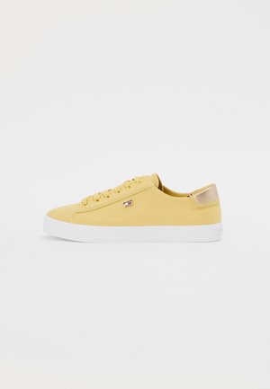 Zapatilla de lona amarilla con suela de goma blanca, punta redonda, seis ojetes y acento en el talón de metal dorado. Pequeño logo en el lateral.