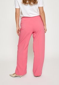 Roza široke hlače z gladko teksturo, opremljene z zadnjimi žepi, v kombinaciji z belo majico in svetlimi čevlji.