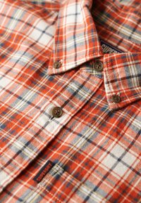 Camicia di flanella a quadri rossa e bianca con accenti blu e gialli, con chiusura a bottoni e etichetta con marchio sul petto.