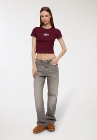 Maglietta cropped bordeaux con la scritta "BROOKLYN NYC" stampata in bianco, abbinata a jeans grigi a gamba larga e scarpe da slip-on marroni. Texture lisce.