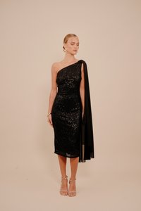 Fekete flitteres ruha aszimmetrikus, egyvállas kialakítással, amelyhez egy folyékony fekete köpeny tartozik; áttetsző sarokkal ellátott szandállal viselve.