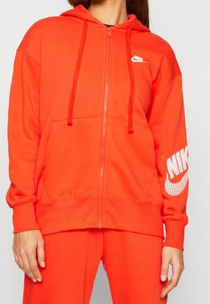 Kobieta ubrana w jasnopomarańczową bluzę Nike na suwak z sznurkami oraz pasujące spodnie, logo Nike na klatce piersiowej i rękawie.