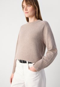 Světle taupe pletený svetr s dlouhými rukávy, kulatým výstřihem, žebrovanými manžetami a jemnými bočními detaily, doplněný bílými kalhotami a černým pásem.